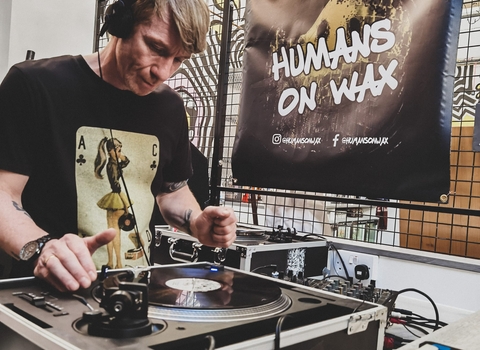 A man with headphones and a playing card t-shirt stand in front of a DJ set, with a Humans On Wax banner in the background