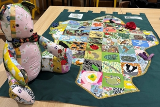 Photo of Lemmy the Leamington Patchwork bear with a collection of fabrics used and the patchwork artwork itself with different wildlife in the area