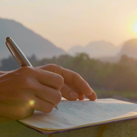 person writing in a notebook with a hill view behind