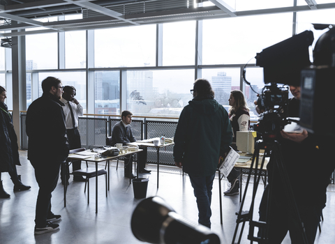 A camera records a group of people in a large windowed office.